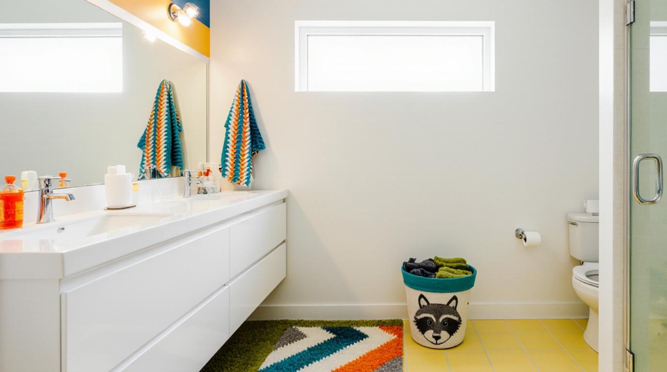 Kids bathroom with white vanity, colorful towels, and a raccoon-themed trash can.