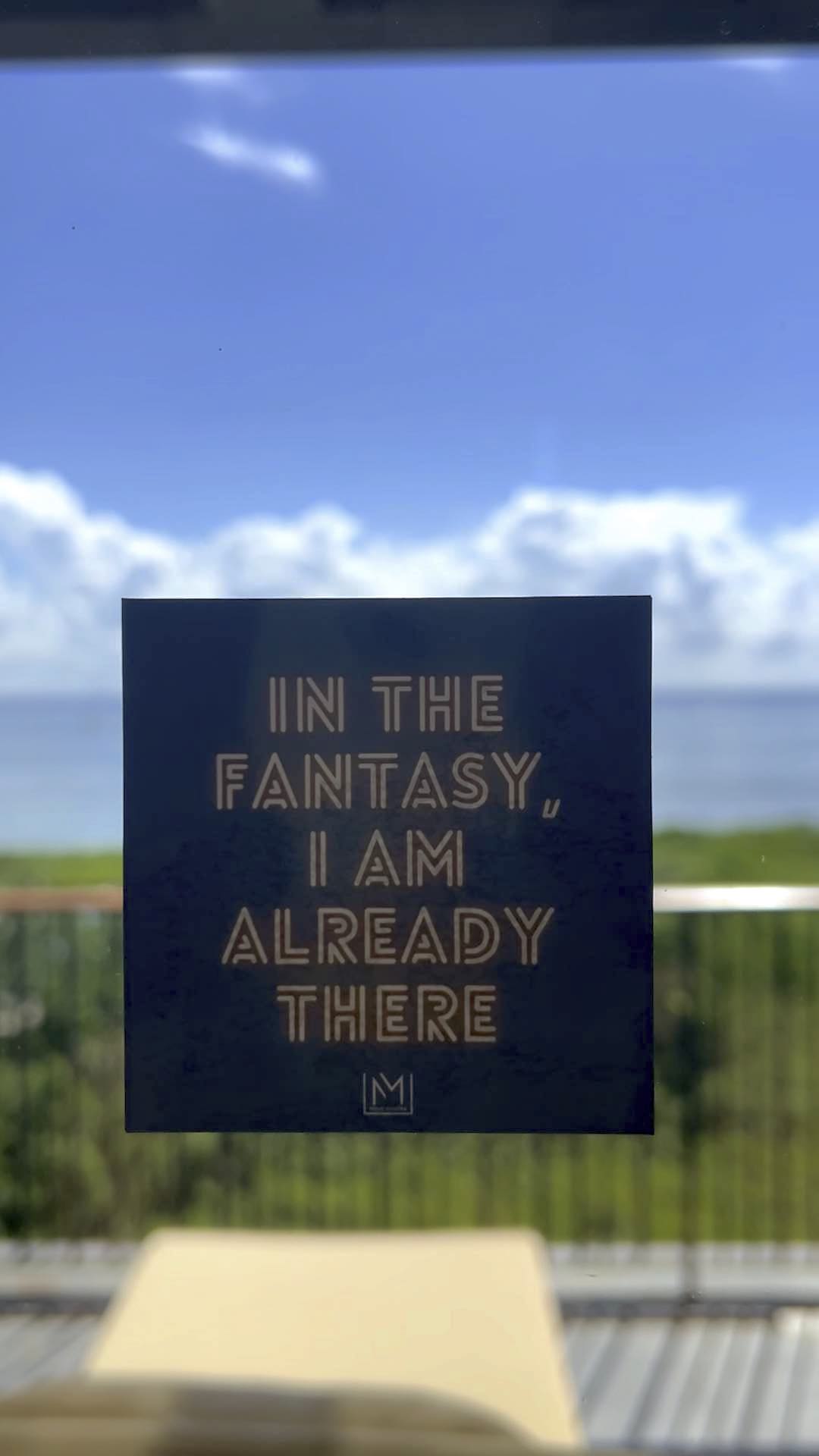 Glass window with a decal applied to it that reads "In the fantasy, I am already there" on a blurred outdoor ocean view balcony background